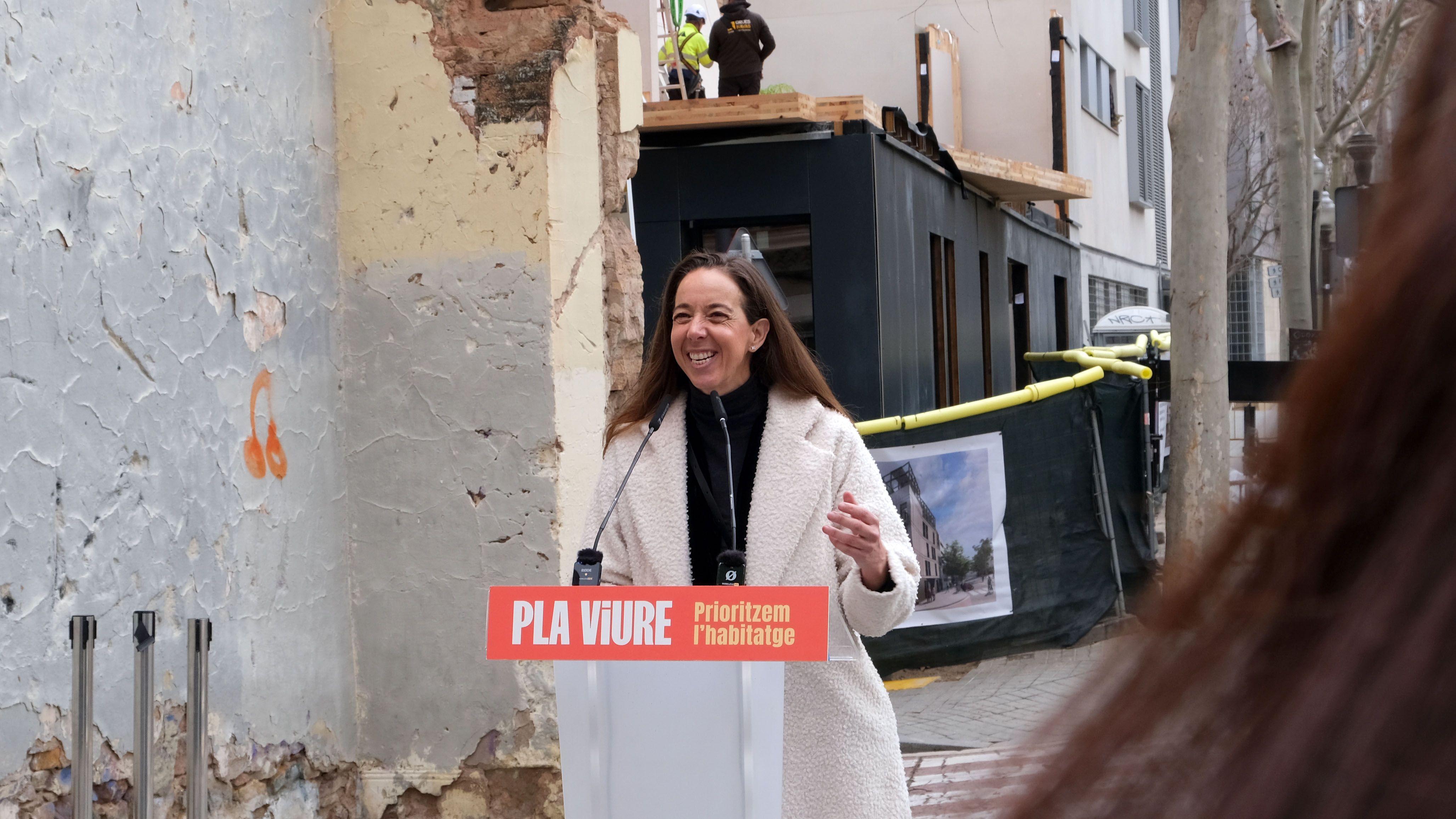 Carlota Pi interviniendo en un acto público frente a un edificio en rehabilitación, con el lema “Pla Viure – Prioritzem l’habitatge”.